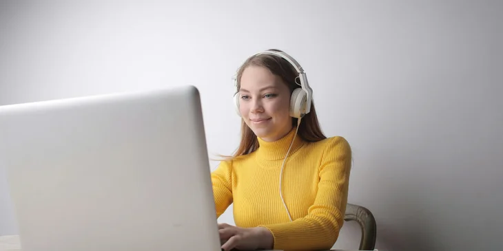 A woman in a yellow sweater wearing headphones while using a laptop.