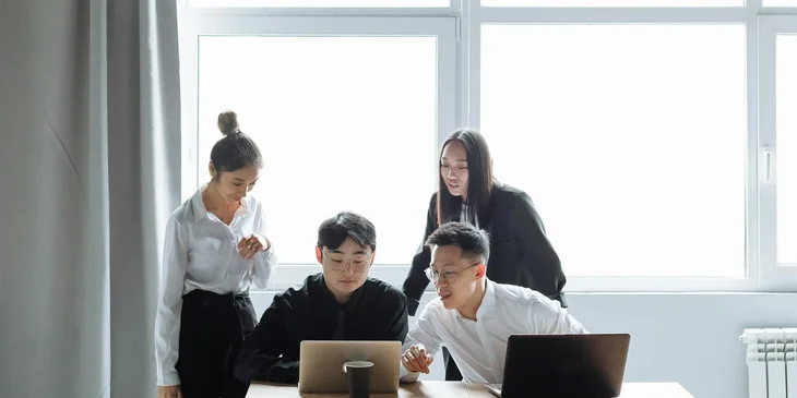 Four students gathered around a desktop and laptops discussing a project.