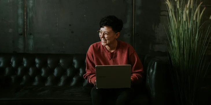 A smiling man using a laptop while sitting on black leather sofa.