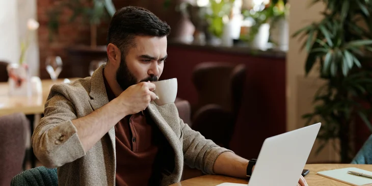 A man sipping coffee while working on a laptop.