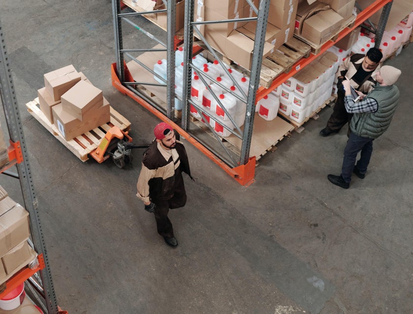 A warehouse sorter moving packages from one spot to another while two co-workers stand nearby discussing work.