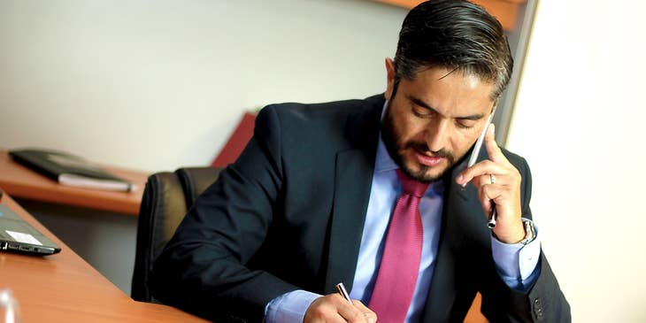 A Business Lawyer in an office reading a book while talking with a client on the phone.