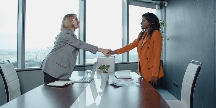 female Customer Relationship Manager wearing gray coat shaking the hands of a female client in orange coat inside the office while smiling