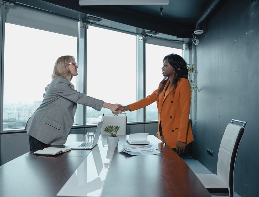 female Customer Relationship Manager wearing gray coat shaking the hands of a female client in orange coat inside the office while smiling