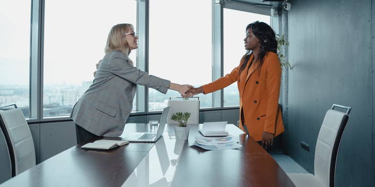 female Customer Relationship Manager wearing gray coat shaking the hands of a female client in orange coat inside the office while smiling