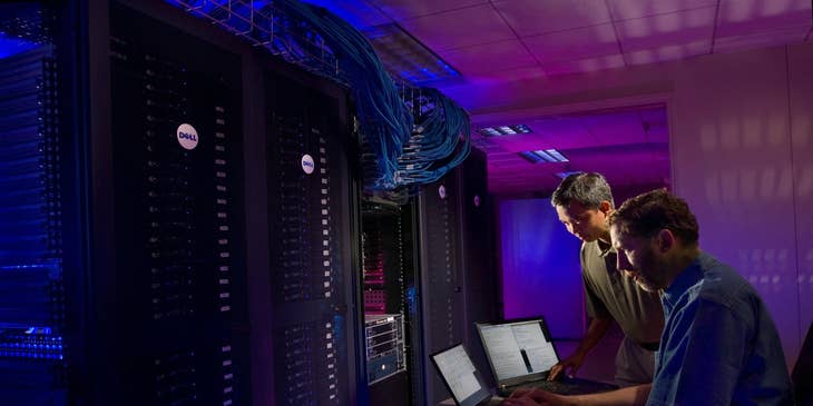 Database Manager working on upgrading the data retrieval system inside the server room while his colleague looks on