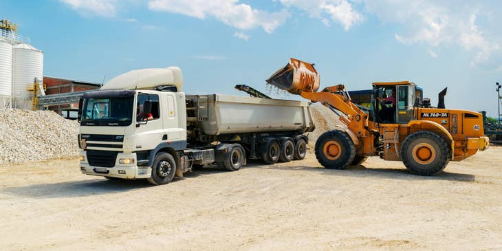 A dump truck driver dumping crushed rocks with the help of construction worker at a construction site.