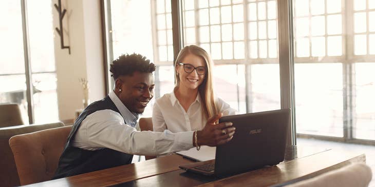 Financial advisor with eyeglasses smiling while looking at a laptop with her male colleague pointing something at the screen