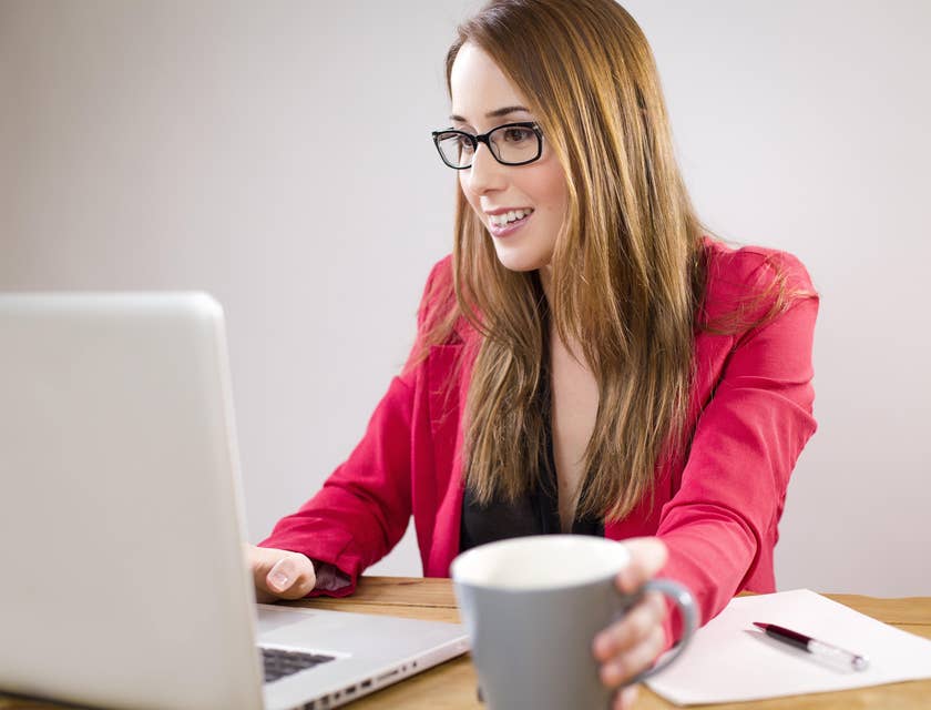 Financial clerk looking at the laptop on her office desk while holding a gray mug