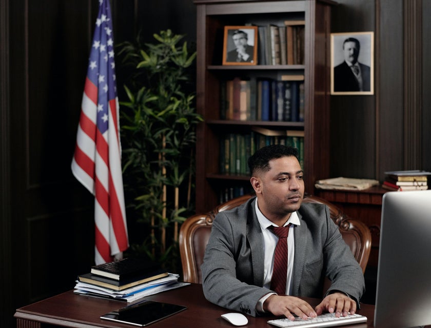 Foreign service officer sitting at his workstation with the bookshelf and the American flag behind him