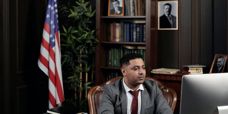 Foreign Service Officer sitting at his workstation with the bookshelf and the American flag behind him