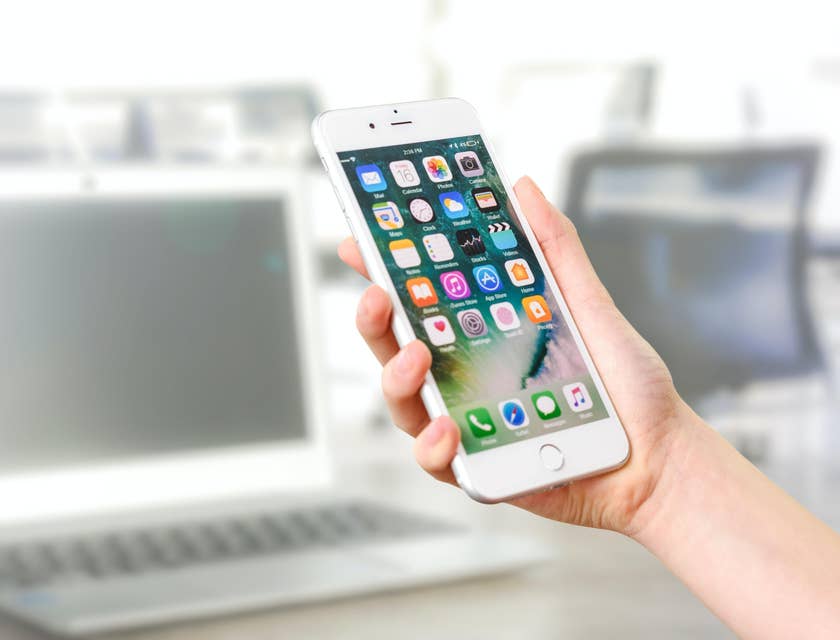 A person holding a phone with app icons and a laptop in the background.