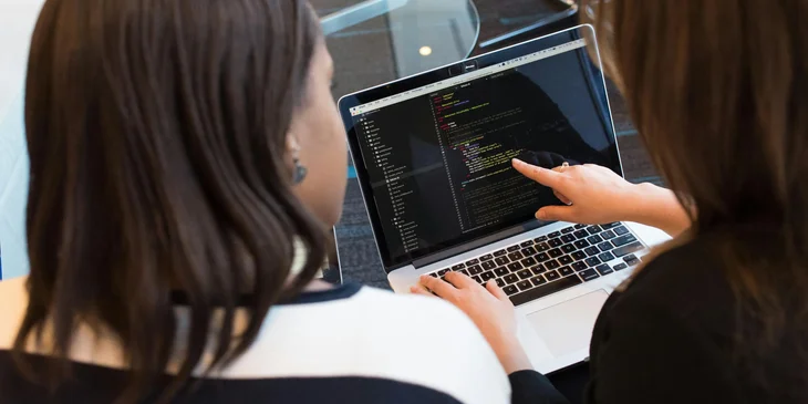 A female HRIS manager working on some codes on a laptop with a coworker.