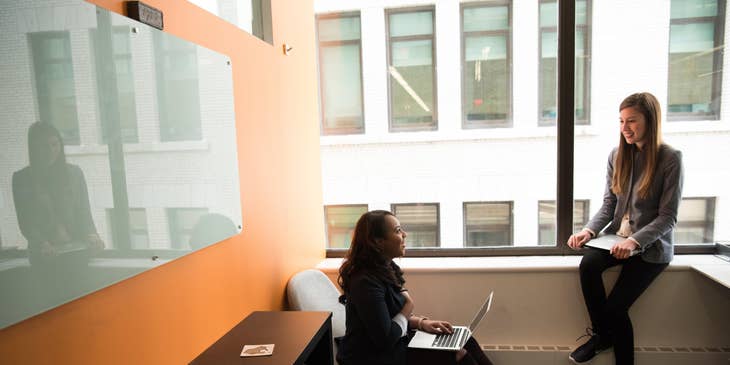 female IT Support Specialist sitting on a chair with her laptop and having discussions with her colleague