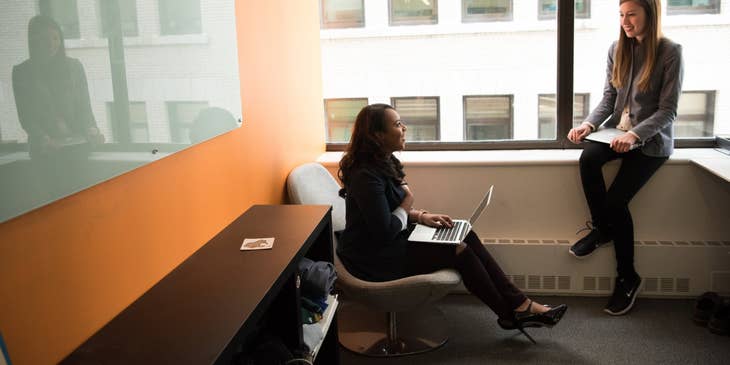 female IT Support Specialist sitting on a chair with her laptop and having discussions with her colleague