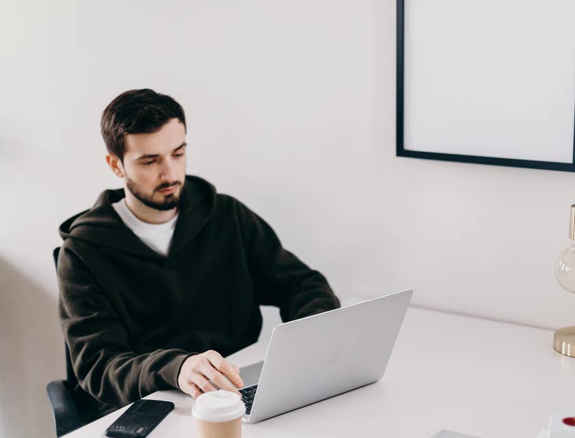 Java software engineer looking at the laptop with his mobile phone and a cup of coffee on the table.