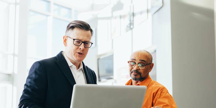 Male life insurance agent in a black suit talking to a client in front of a laptop.