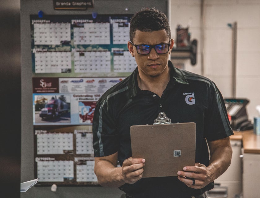 A logistics associate holding a clipboard in an office warehouse reviewing shipment paperworks.