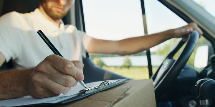 A Long Haul Truck Driver doing inventory on an inventory sheet on a clipboard on top of a box inside a truck.