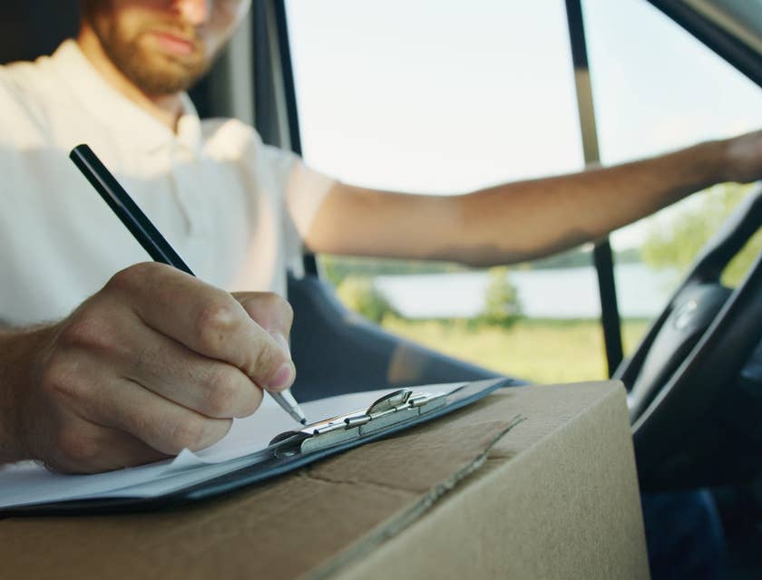 A long haul truck driver doing inventory on an inventory sheet on a clipboard on top of a box inside a truck.