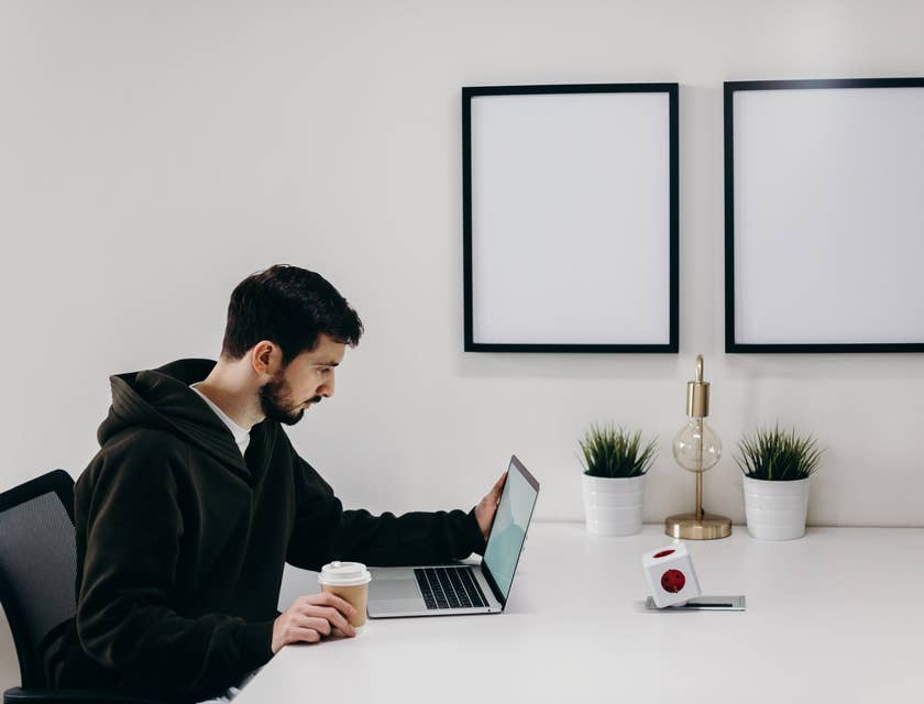 Magento developer sitting at a white table and looking at his laptop while holding a cup of coffee