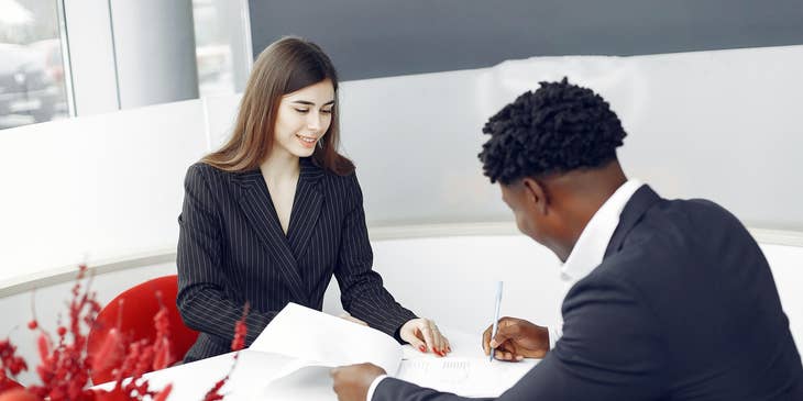Female mortgage processor smiling and looking over a client signing papers on the table.