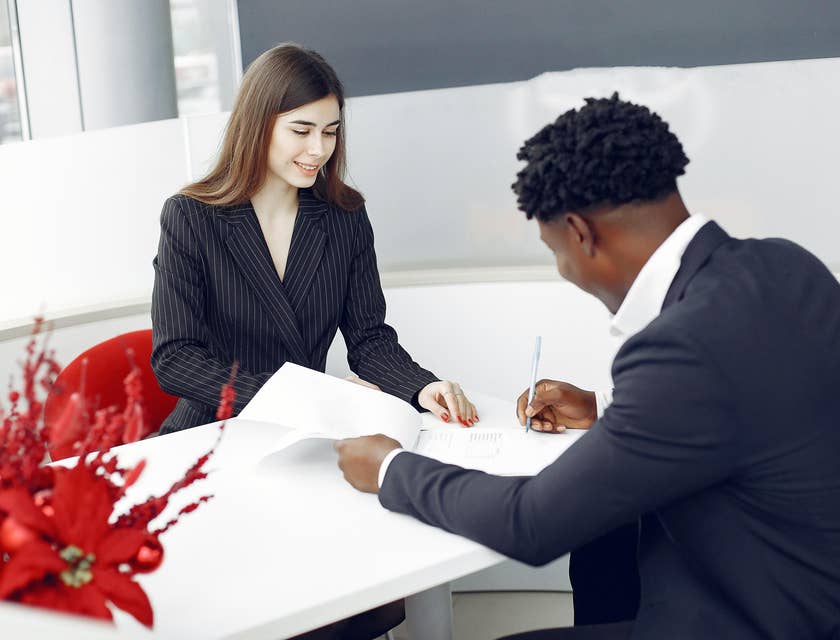 Female mortgage processor smiling and looking over a client signing papers on the table.
