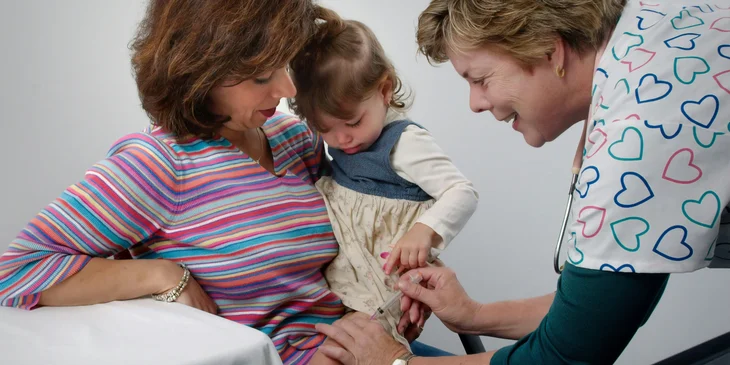 A pediatric medical assistant gives vaccine shot on the leg as the patient's mother comforts her.