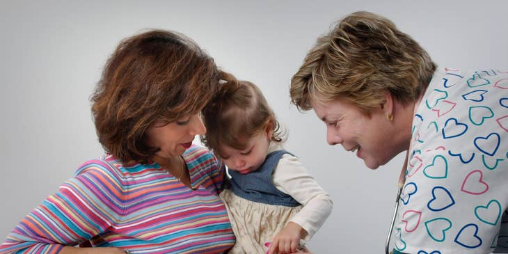 Pediatric medical assistant gives vaccine shot on the leg as the patient's mother comforts her.