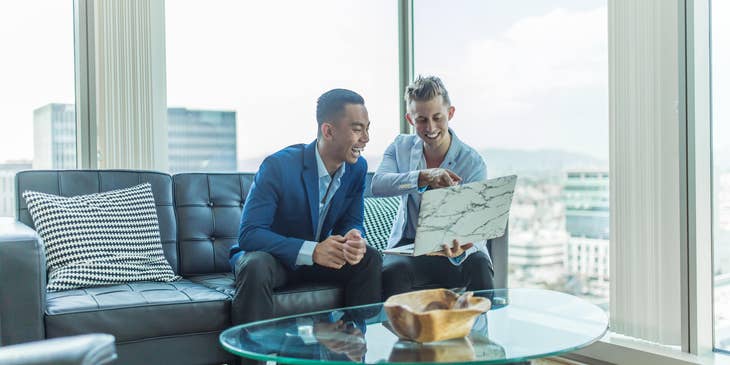 Young personal banker showing details at his open laptop to his client while sitting on a couch.