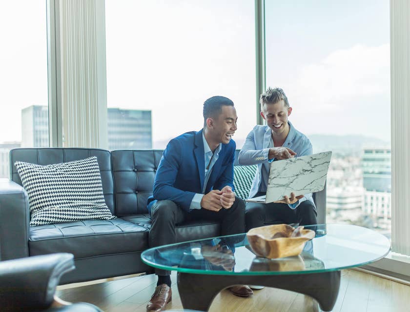 Young personal banker showing details at his open laptop to his client while sitting on a couch.