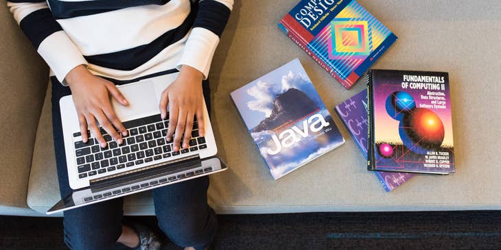 React.js developer sitting on a couch with computing books while typing on a laptop
