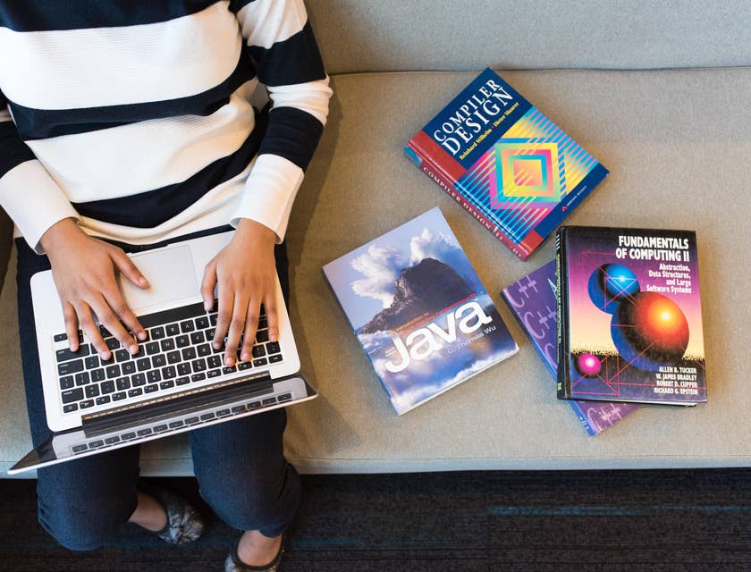 React.js developer sitting on a couch with computing books while typing on a laptop