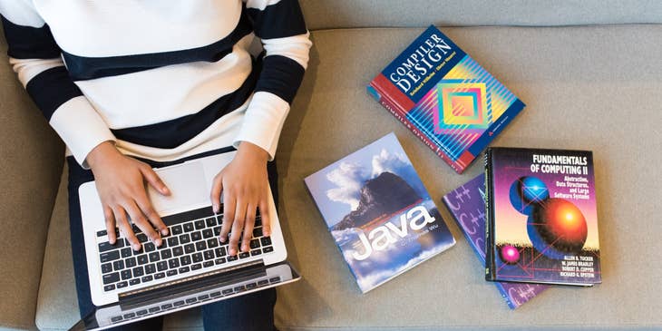 React.js Developer sitting on a couch with computing books while typing on a laptop