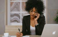 Uma mulher trabalhando em um laptop, fazendo anotações e falando ao celular.