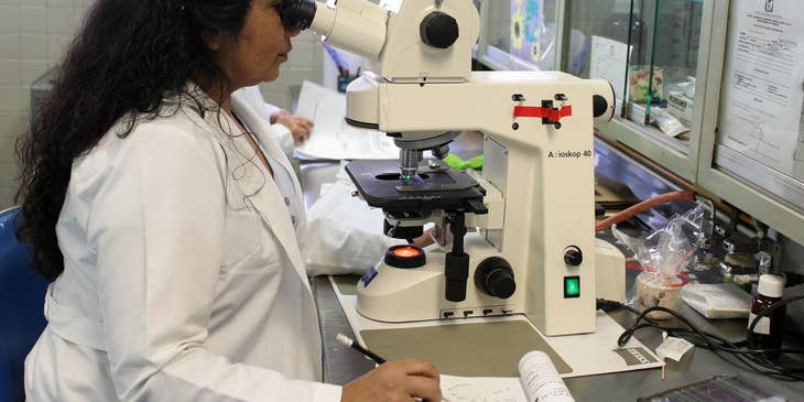 Research scientist looking down a microscope and writing down her observations.
