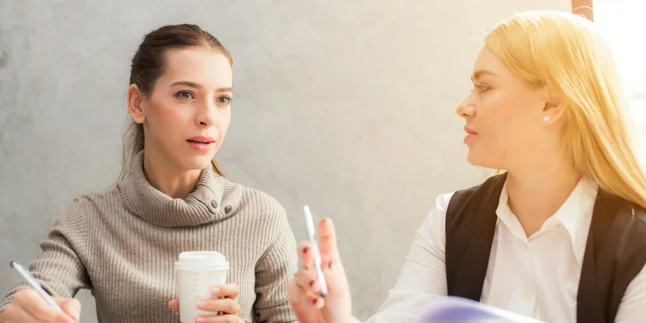 A retention specialist communicates with sales rep discussing strategies while taking notes and drinking coffee.