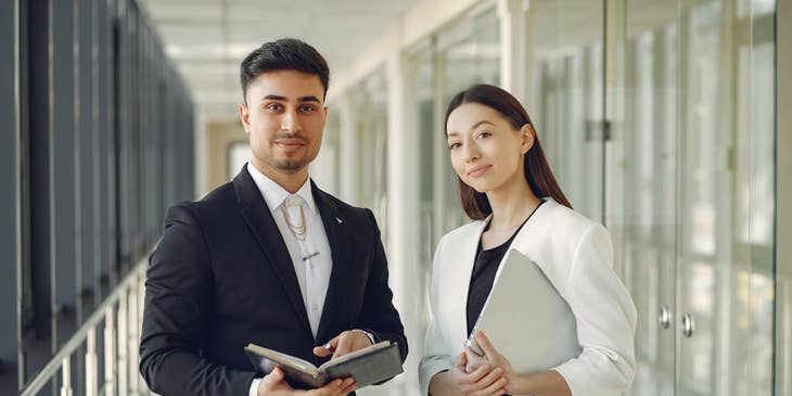 two senior financial analysts holding a notebook and laptop while standing in the hallway