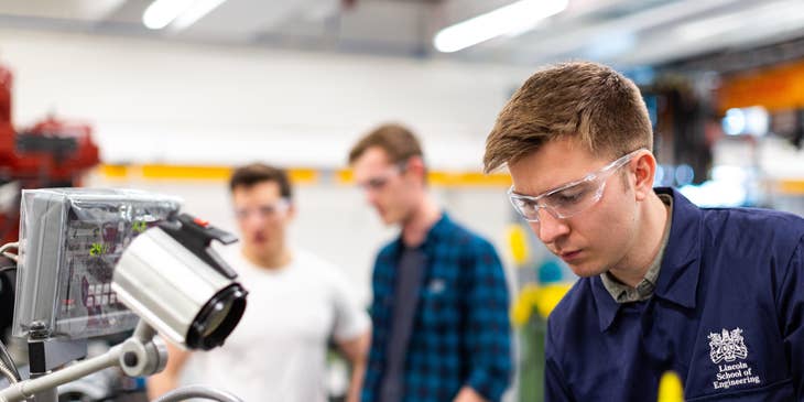Staff engineer wearing protective goggles while putting together the manufactured product in a laboratory with his colleagues on the background