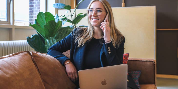 female transfer agent wearing eyeglasses and sitting on the couch with her open laptop while using her mobile phone