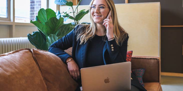 female Transfer Agent wearing eyeglasses and sitting on the couch with her open laptop while using her mobile phone