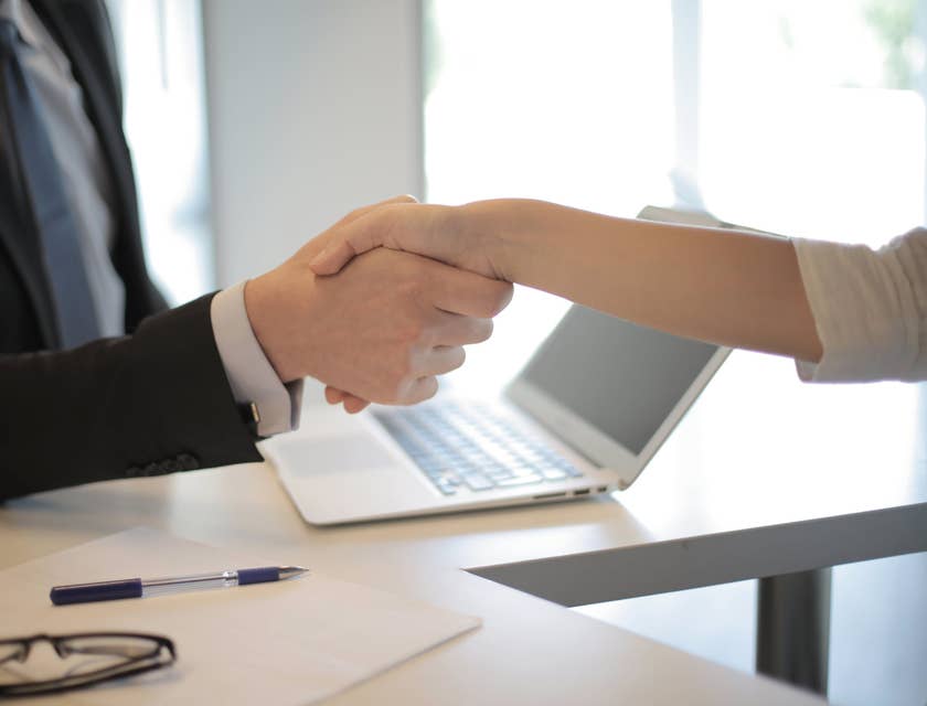 Wealth Manager wearing a business suit shaking hands with a client over an open laptop, eyeglasses, piece of white paper and a pen